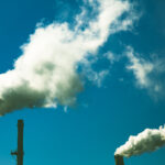 The image shows two industrial chimneys polluting the blue sky with thick, white plumes of smoke.